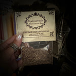 A hand holds a small clear pouch containing dried brown herbal material, with a cream label reading 'Bladderwrack Herb of the Sea' and 'Smudge Metaphysical.' The scene appears to be a retail or display setting with other jars and dried botanicals in the background.