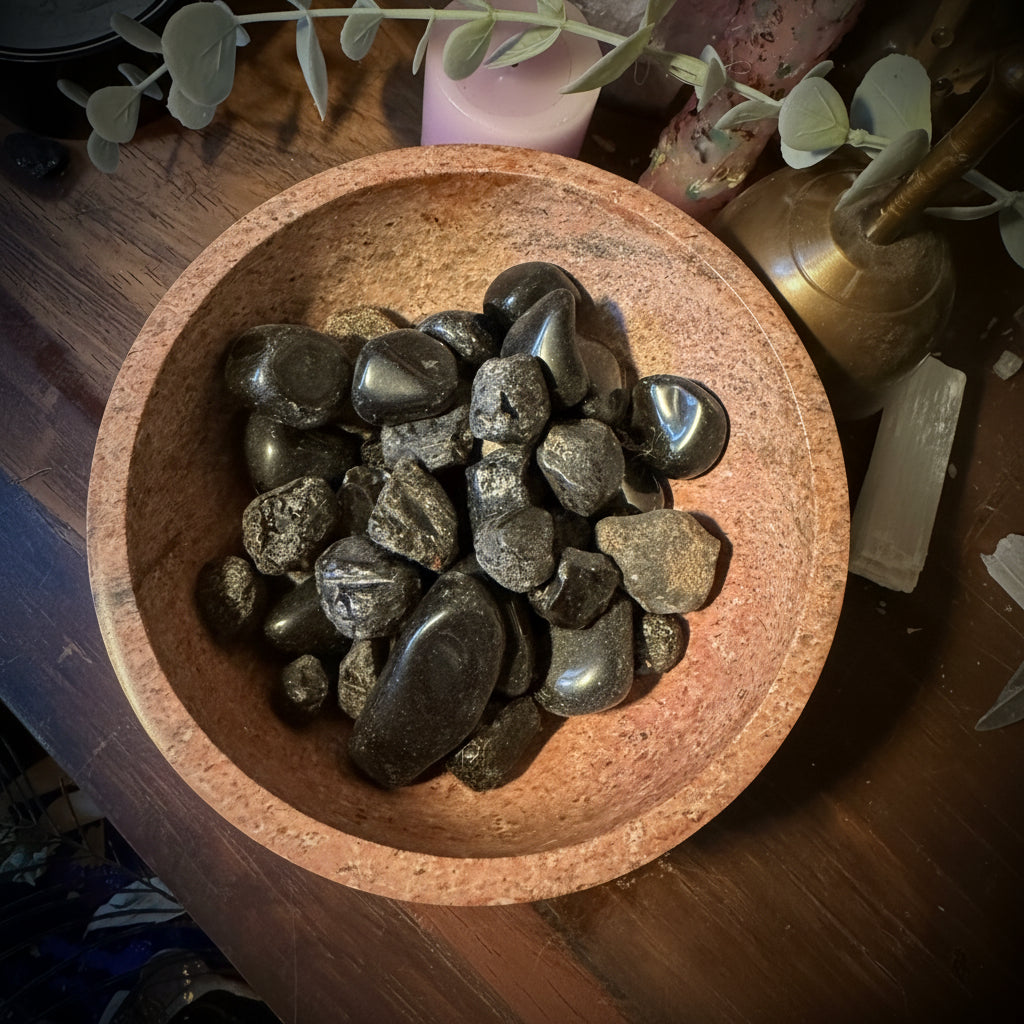 A bowl filled with polished black tumbled stones, photographed on a wooden surface with crystals and decorative props around it.