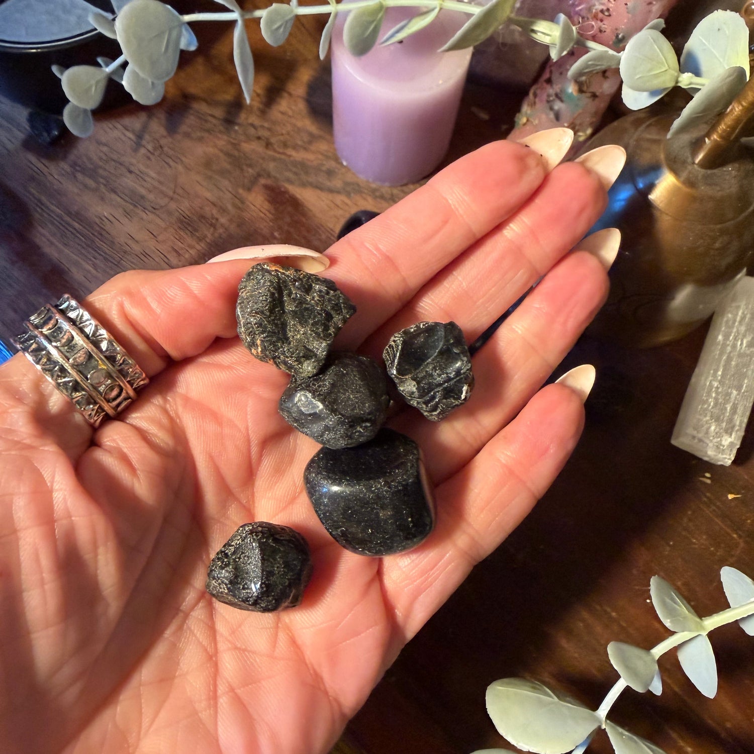 A hand holding five polished black tumbled stones, with a wooden table, candles, crystals, and eucalyptus branches in the background.