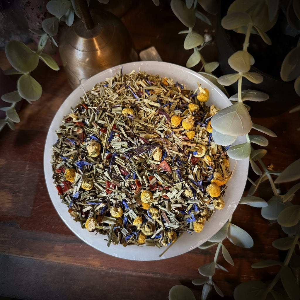 A white bowl filled with loose-leaf herbal tea, featuring dried flowers, green herbs, and small yellow berries, styled on a wooden surface with eucalyptus leaves and a brass candle holder.