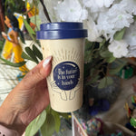 A hand holding a reusable coffee cup with a navy blue lid and a navy blue circular graphic that reads 'The future is in your bands' with a moon and stars design. The cup body is a warm cream/beige color.