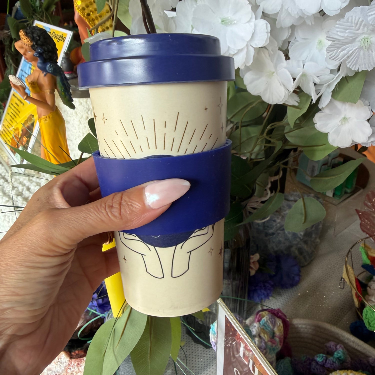 A hand holding a reusable coffee cup with a dark blue lid and a matching blue sleeve around the middle. The cup body is a warm cream color with a simple line drawing of hands and rays.