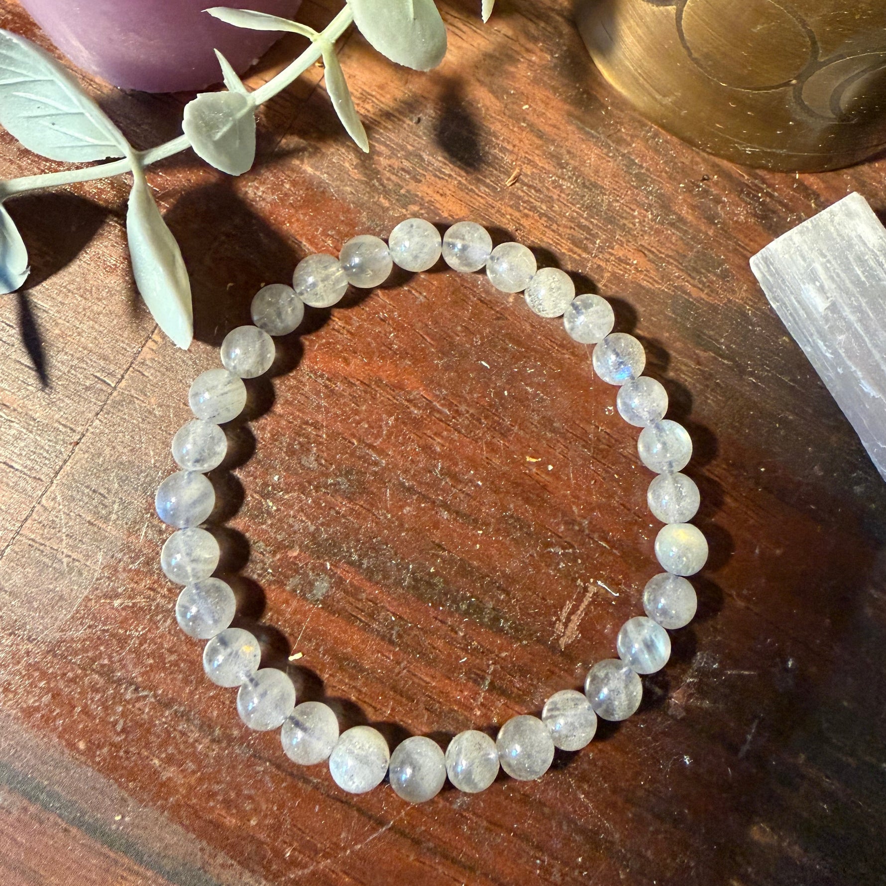 A beaded stretch bracelet made of round, pale grayish-blue stones, displayed on a dark wooden surface with decorative props including a purple candle, green leaves, a brass object, and a white crystal.