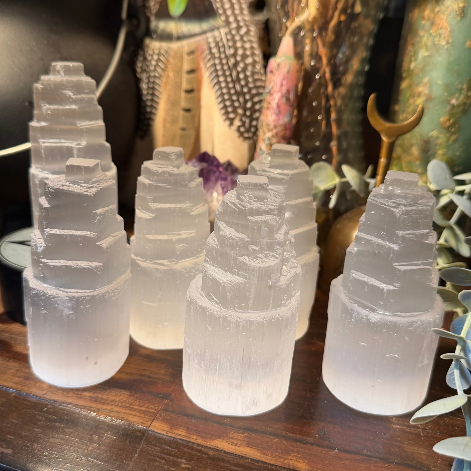 Five white selenite tower lamps arranged on a dark wooden table in a styled home decor setting with dried flowers, a cauldron, and greenery.