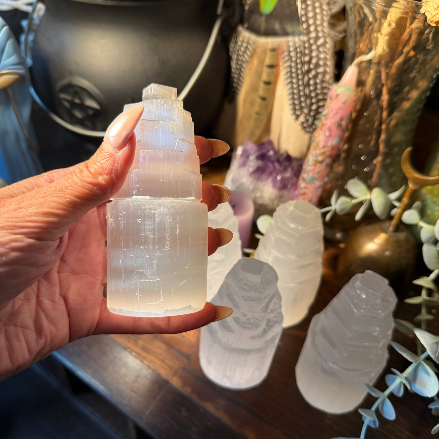 A hand holds a frosted white selenite tower crystal against a dark, decorated background with other crystal formations on a wooden surface.