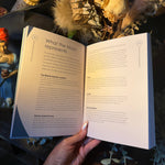 A hand holds an open book showing printed pages about what the moon represents, with dried flowers and decorative objects in the background.