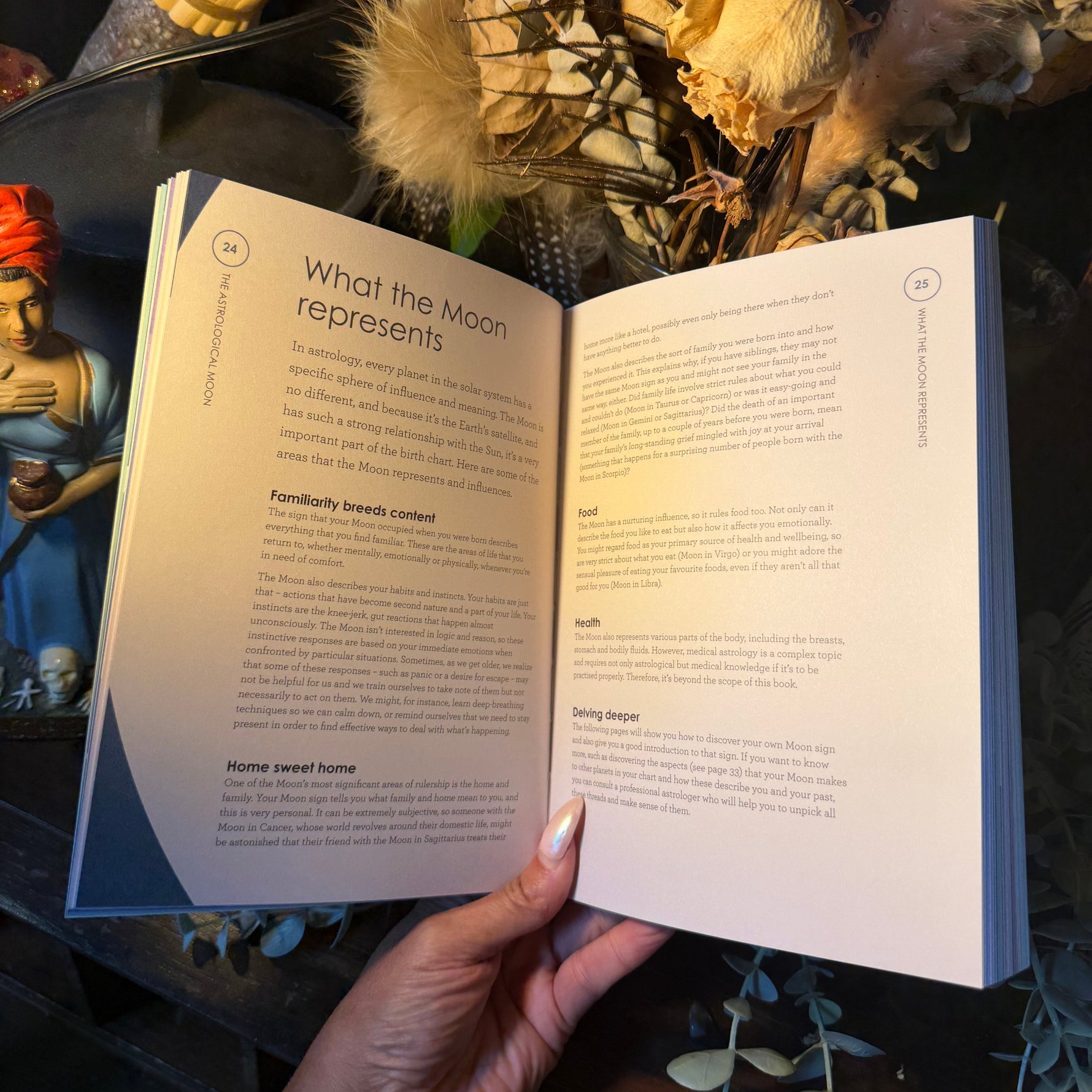A hand holds an open book showing printed pages about what the moon represents, with dried flowers and decorative objects in the background.