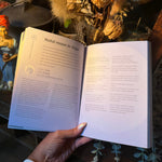 An open book titled 'Natal moon in Vagi' being held by a hand, set against a background of dried flowers and decorative figurines.