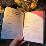 An open booklet or planner showing pages about work and financial management, held in a hand against a styled background with dried flowers and decorative figurines.