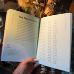 An open book showing printed text about 'The Moon's nodes' held by a hand, with dried flowers and greenery arranged around it on a wooden surface.