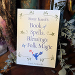 A book titled 'Sister Karol's Book of Spells, Blessings & Folk Magic' by Karol Jackowski, standing upright on a wooden surface surrounded by dried flowers, greenery, and candles.