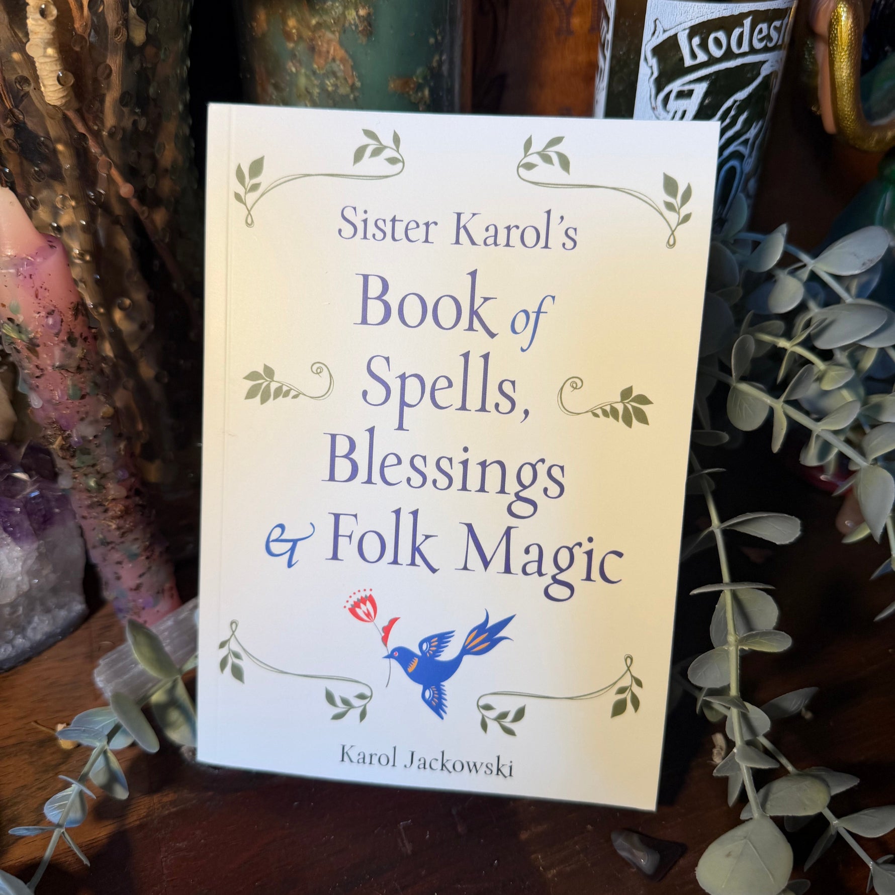 A book titled 'Sister Karol's Book of Spells, Blessings & Folk Magic' by Karol Jackowski, standing upright on a wooden surface surrounded by dried flowers, greenery, and candles.