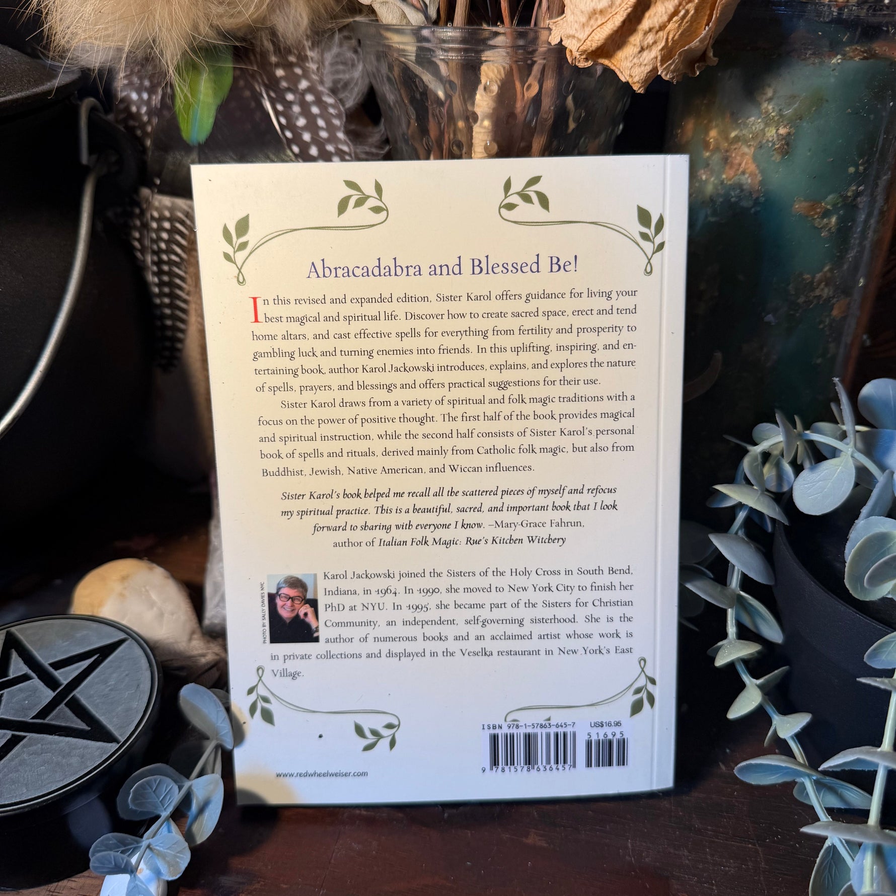 The back cover of a book titled 'Abracadabra and Blessed Be!' is displayed on a rustic wooden surface surrounded by mystical props including a black pentacle, dried flowers, and eucalyptus leaves.