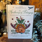 The Apothecary of Belonging book by Alexis J. Cunningfolk, featuring a detailed illustration of a winged heart with flowers and leaves on the cover, displayed on a wooden surface surrounded by dried botanicals and eucalyptus.