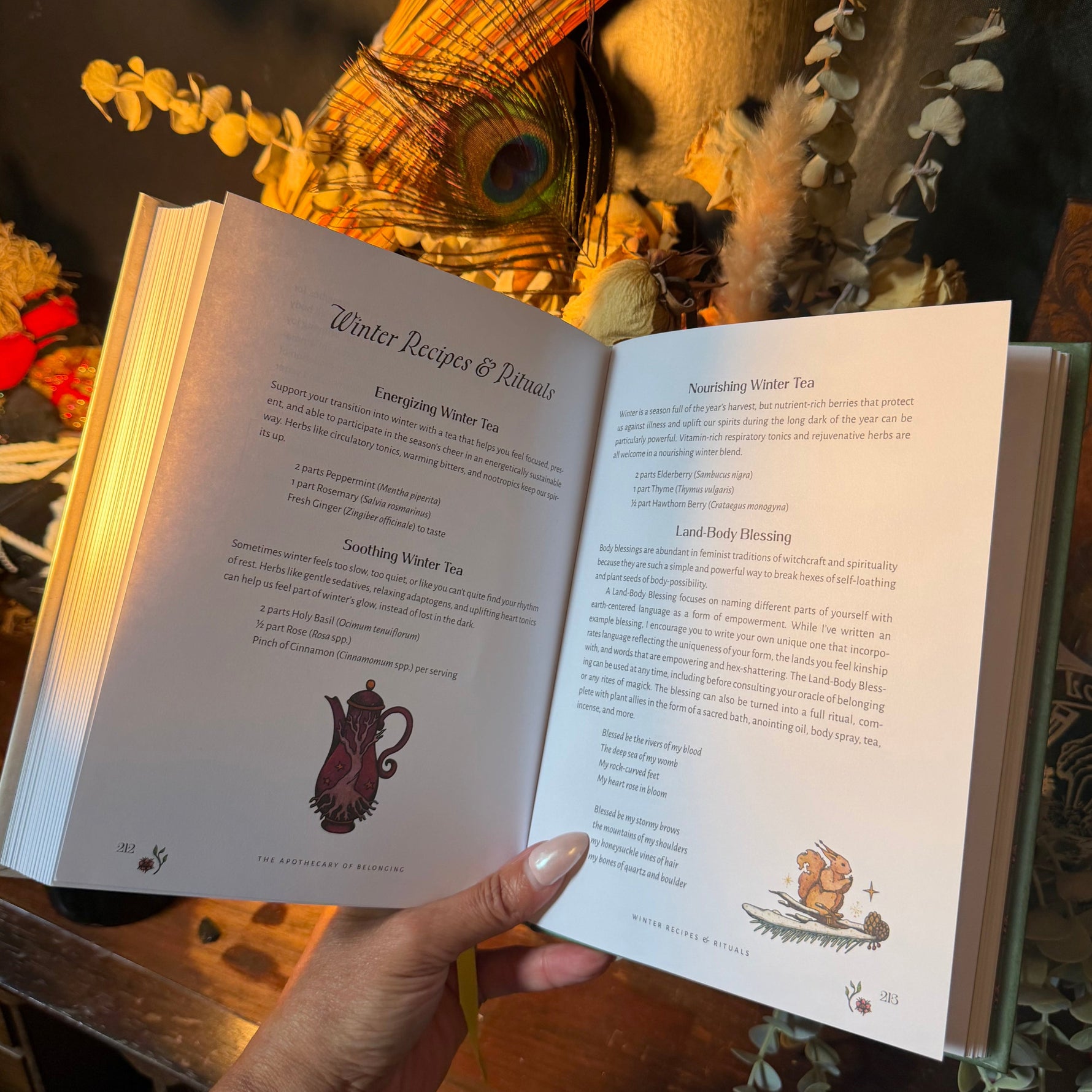 A hand holds an open book showing printed recipe pages, with a warm decorative background featuring dried flowers, a peacock feather, and a wooden surface.