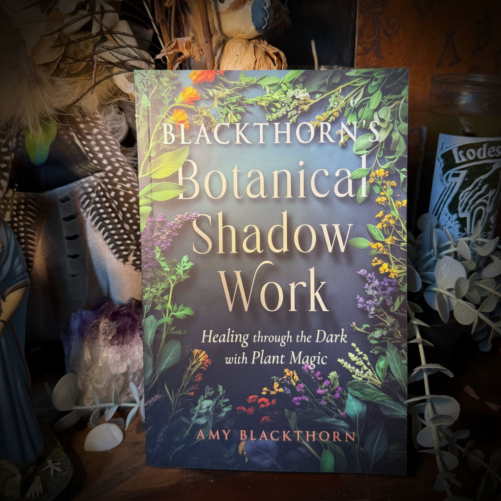 The book 'Blackthorn's Botanical Shadow Work' by Amy Blackthorn is displayed on a wooden surface surrounded by mystical props including dried flowers, a Lodestar jar, and crystals.