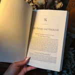 A hand holds an open book showing a printed page titled "Talk Therapy and Witchcraft," surrounded by dried flowers, greenery, and decorative objects on a dark surface.