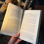 An open book held by a hand, showing printed text pages, with decorative dried grasses, a peacock feather, and eucalyptus leaves in the background.
