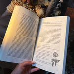 An open book held by a hand, showing printed text and a botanical illustration on the right page, with dried flowers and a peacock feather in the background.