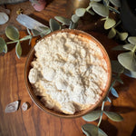 A rustic brown ceramic bowl filled with a textured, crumbly off-white powder, set on a wooden table surrounded by green eucalyptus leaves and crystals.