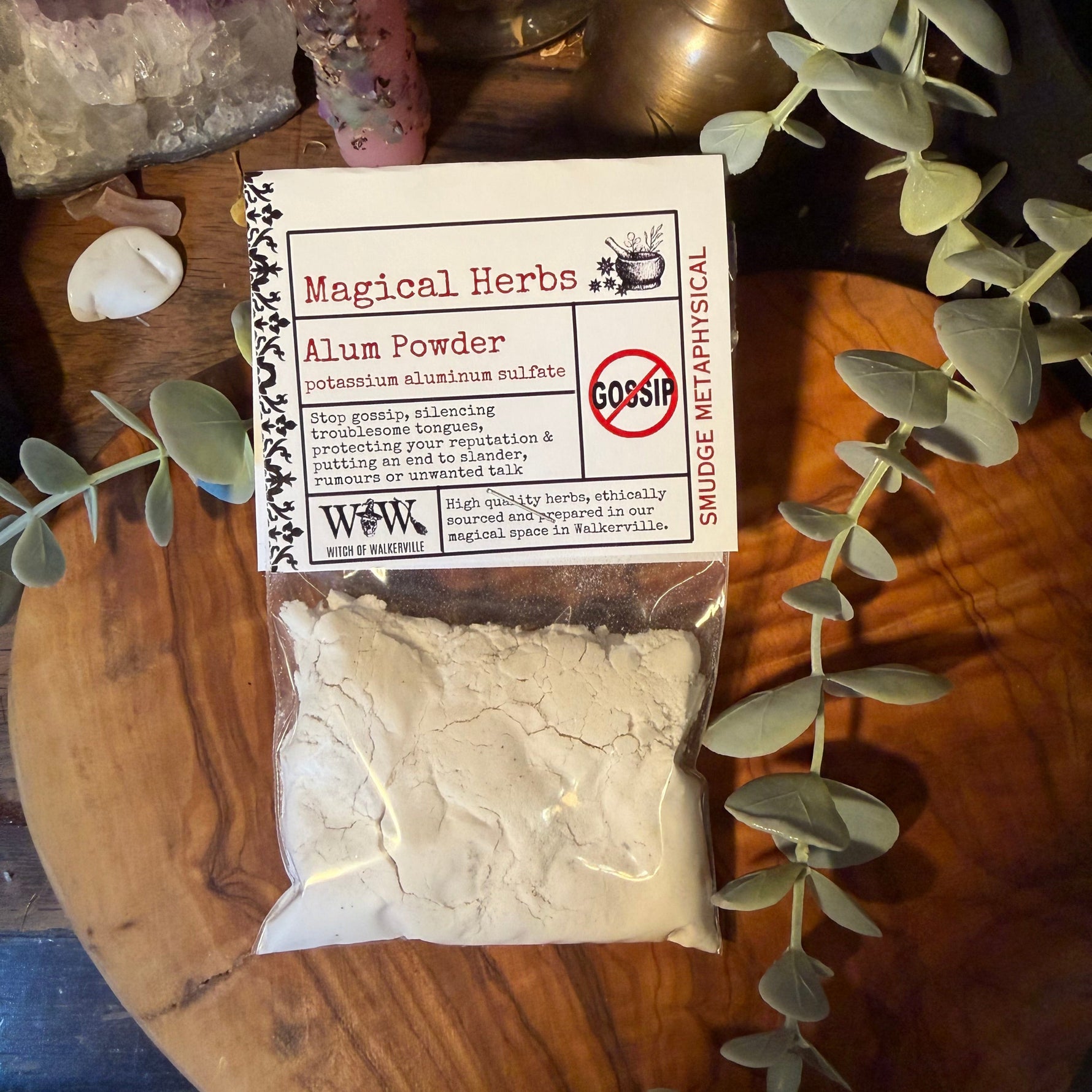 A clear plastic bag filled with white aluminum powder sits on a round wooden board, with a printed label reading 'Magical Herbs Alum Powder'  Decorative greenery and crystals surround the product in a styled setting.