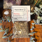 A clear plastic bag filled with dried vervain herb pieces sits on a wooden surface, with a white product label reading 'Magical Herbs' and 'Vervain' placed above it. The scene is styled with eucalyptus leaves, crystals, and decorative objects in the background.