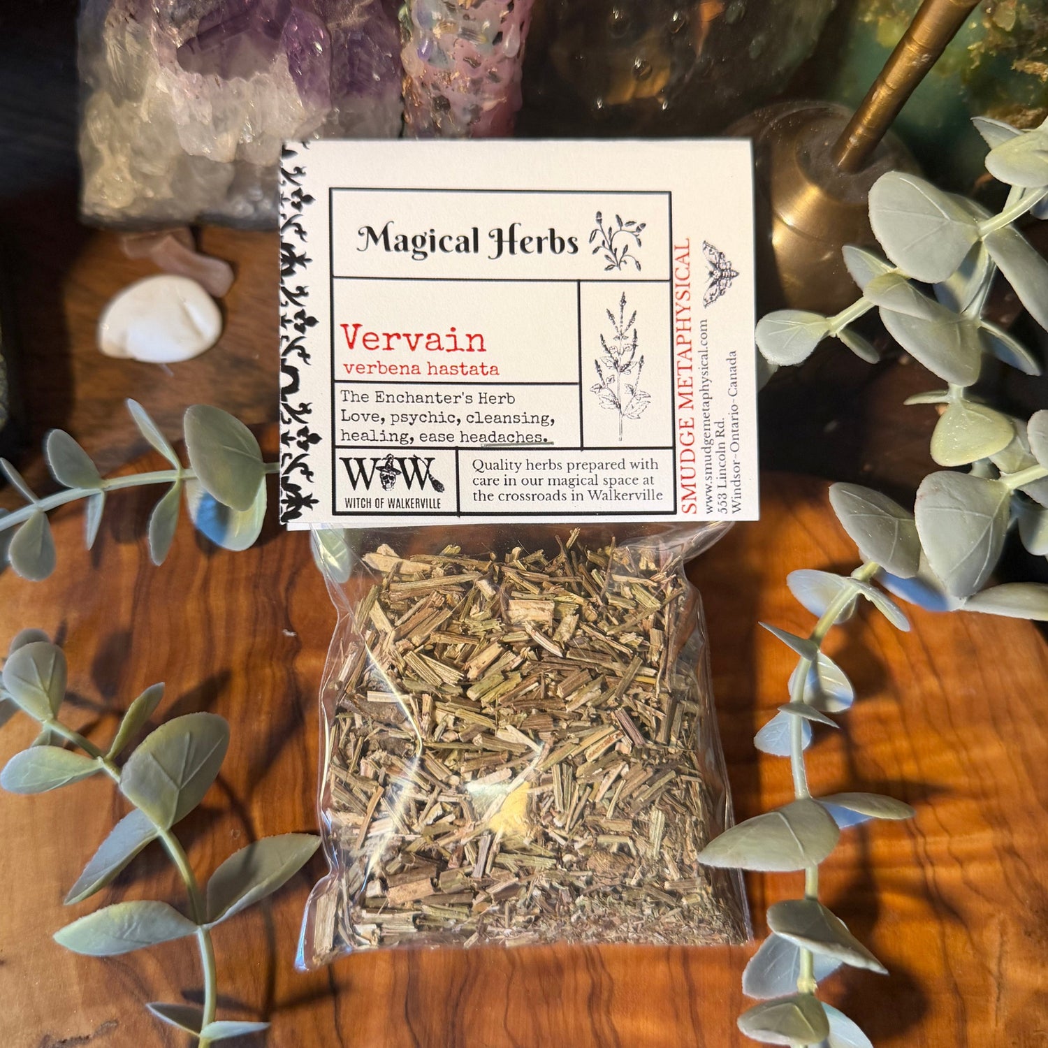 A clear plastic bag filled with dried vervain herb pieces sits on a wooden surface, with a white product label reading 'Magical Herbs' and 'Vervain' placed above it. The scene is styled with eucalyptus leaves, crystals, and decorative objects in the background.