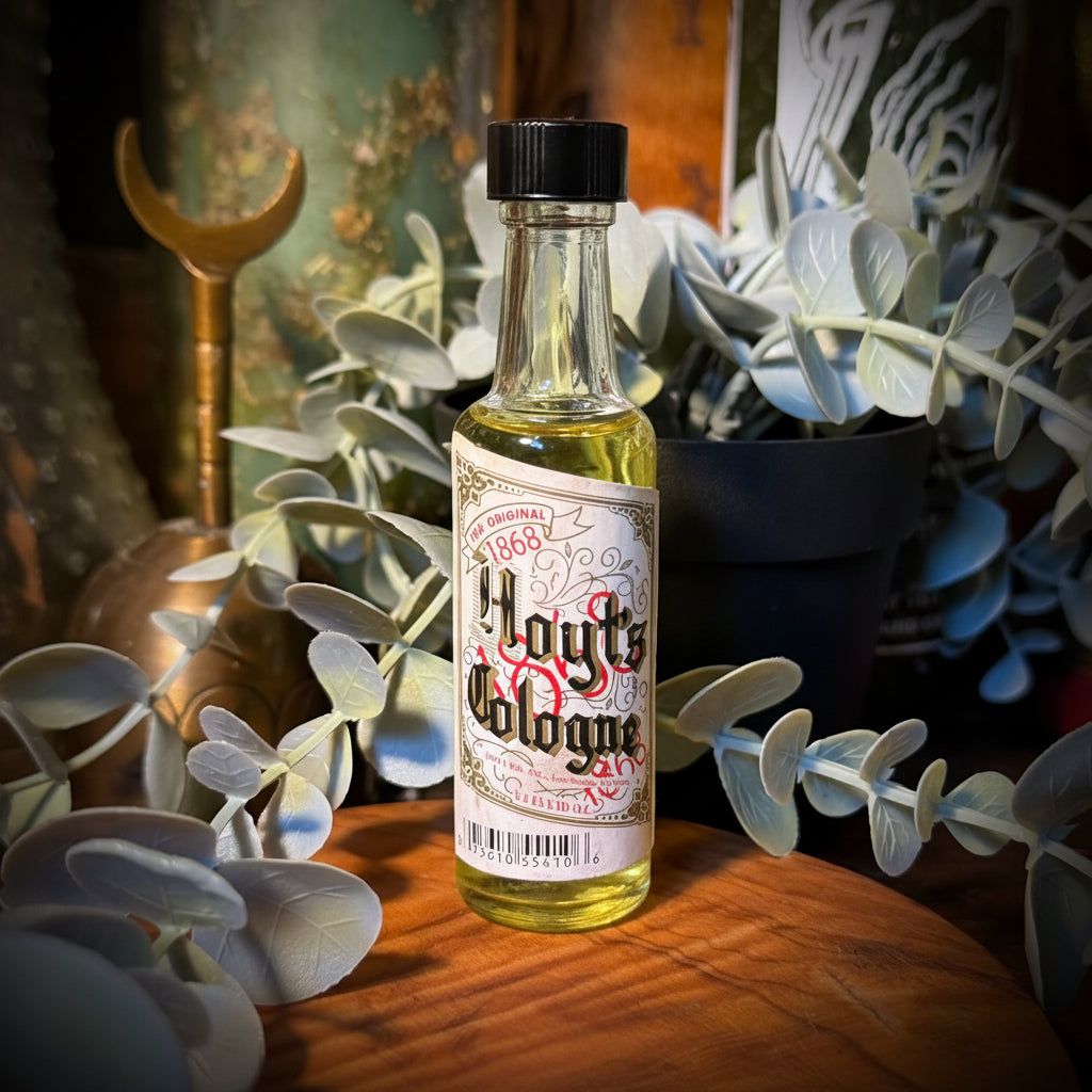 A small glass bottle of Haut's Cologne with a black cap and a vintage-style cream label, placed on a wooden surface surrounded by eucalyptus leaves and decorative objects.