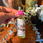 A hand with pink nail polish holds a small bottle of Houghton's Cologne against a rustic background with a wooden surface, dried plants, and eucalyptus.