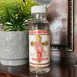 A bottle of Agua Bendita Holy Water standing on a dark wooden surface next to a potted green plant.