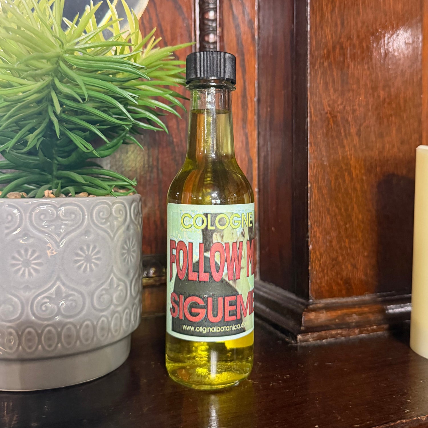 A small glass bottle of 'Follow Me' hot sauce by Ignacio Rodriguez sits on a dark wooden mantelpiece next to a potted green plant and a white pillar candle.
