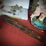 A dark brown wooden wand rest with gold star inlays, shown on a red textured surface next to a book titled 'Year of the Witch' and a small fairy figurine.