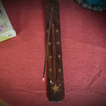 A dark brown wooden incense holder with gold star inlays, containing a single lit incense stick, placed on a textured red tablecloth with books and a decorative figurine in the background.