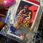 A hand with pink nail polish holds a clear plastic bag containing a small amulet set. The set includes a printed card with an illustrated figure, a red cloth, a gold-tone key, several beads, and a blue circular charm.
