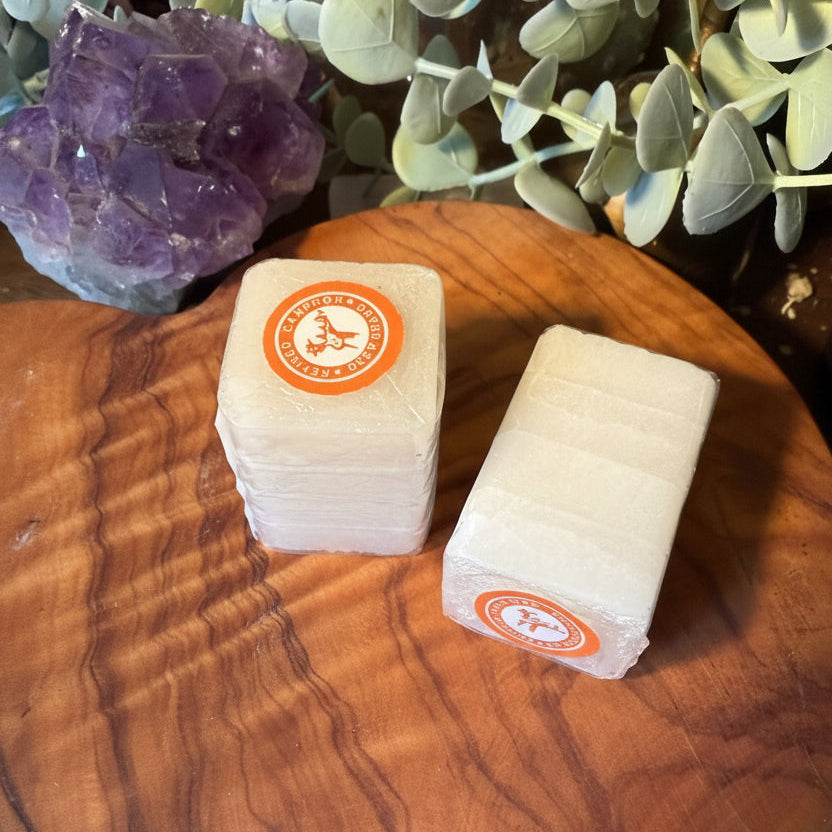 Two rectangular bars of soap wrapped in clear plastic with an orange circular logo, displayed on a round wooden board with eucalyptus leaves and an amethyst crystal in the background.