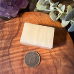 A rectangular bar of pale cream-colored soap sits on a round wooden surface, with a coin placed below it for scale. The scene is styled with eucalyptus leaves and a purple crystal cluster in the background.