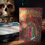 A decorative book titled 'Santa Muerte Book of the Dead' stands on a wooden surface, surrounded by gothic props including a skull with red eye sockets, a stack of books, and a dark vase with pink flowers.