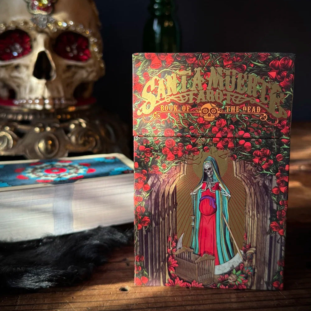 A decorative book titled 'Santa Muerte Book of the Dead' stands on a wooden surface, surrounded by gothic props including a skull with red eye sockets, a stack of books, and a dark vase with pink flowers.