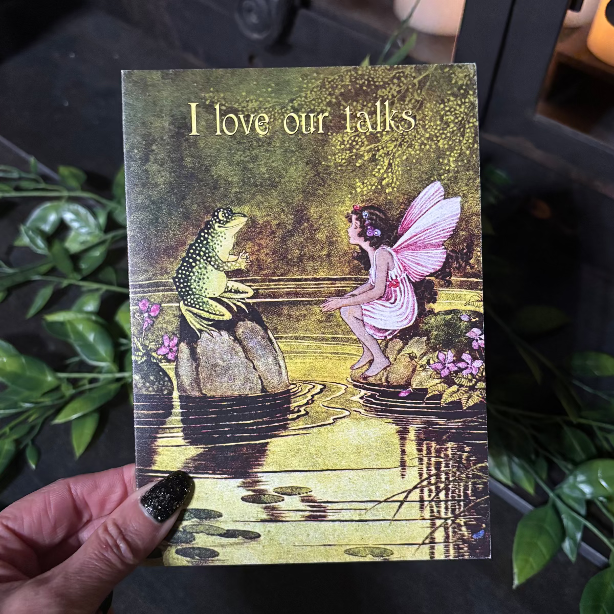 Fairy and frog illustration on a card held by a hand with black nail polish.