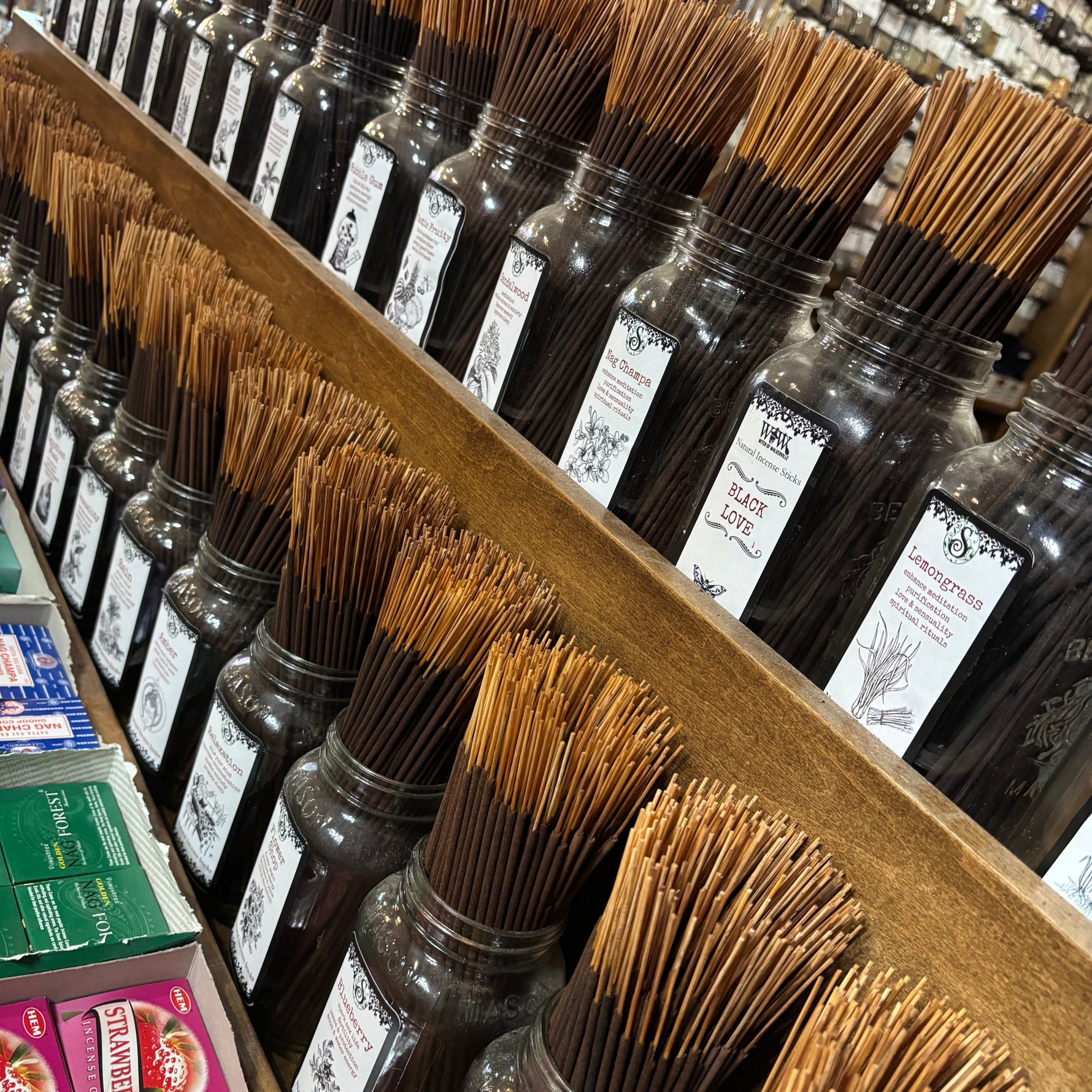 Rows of incense sticks in clear glass jars with white labels, arranged on wooden shelves in a store setting.