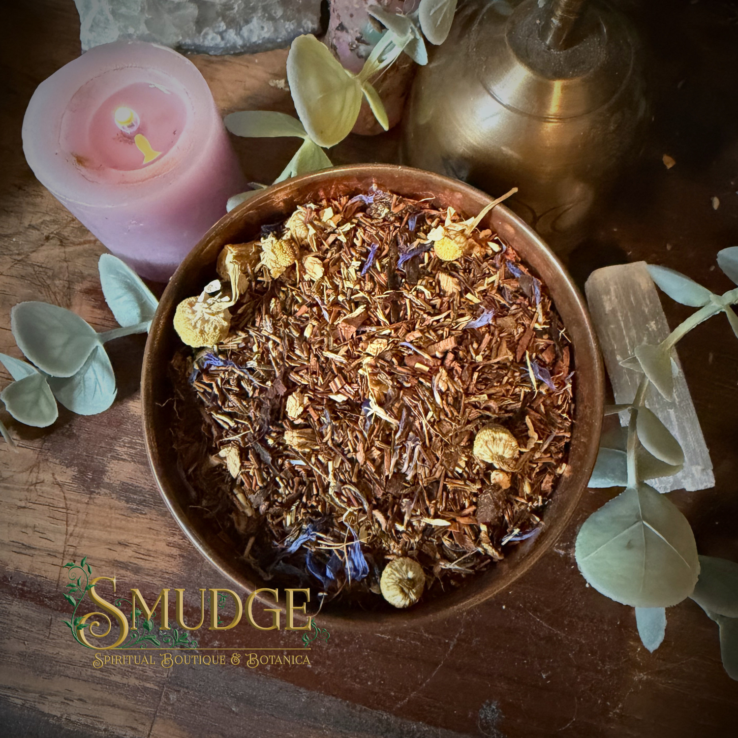 A bowl of loose-leaf spiritual and botanical tea blend from Smudge Spiritual Botanicals, styled on a rustic wooden table with a pink candle, a brass chalice, and green leaves.