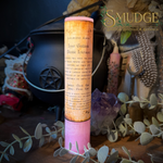 A pink and tan cylindrical spiritual candle labeled 'Liber Benz Aven Upper Goddess Decèssé Interière' stands on a wooden surface surrounded by dried greenery, crystals, and decorative objects. The brand name 'SMUDGE' is visible in the upper right corner.