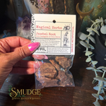 A hand with pink nail polish holds a small clear plastic bag containing dark brown Jezebel Root herbal pieces, attached to a white paper header card labeled 'Magical Herbs' and 'Jezebel Root'. The background is a dark wooden surface with decorative plants and crystals.
