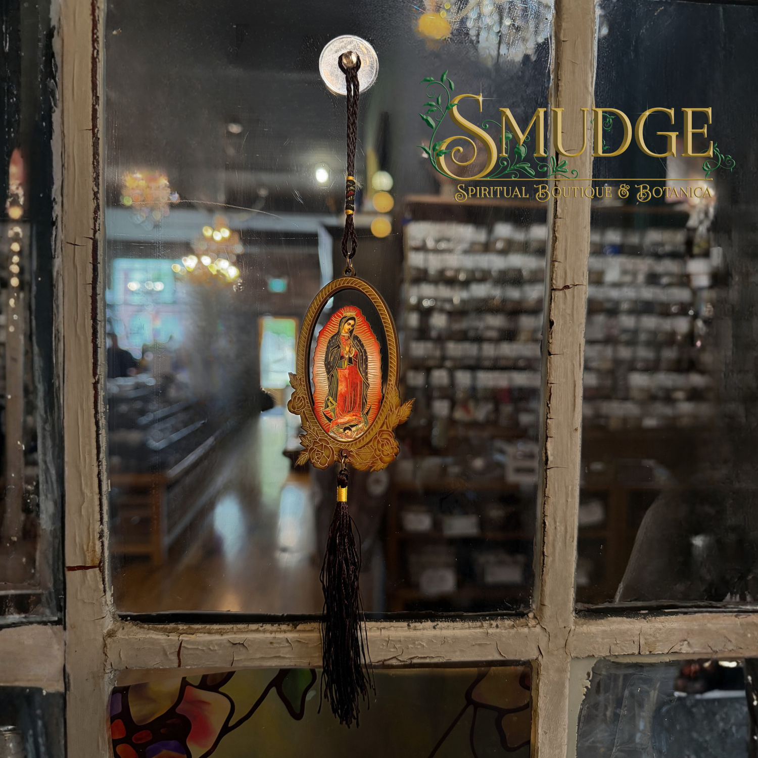 A religious hanging ornament featuring a small oval image of Our Lady of Guadalupe with gold-toned decorative details and a long black tassel, suspended in front of a shop window. The window has the words 'SMUDGE Spiritual Boutique & Botany' printed on it.