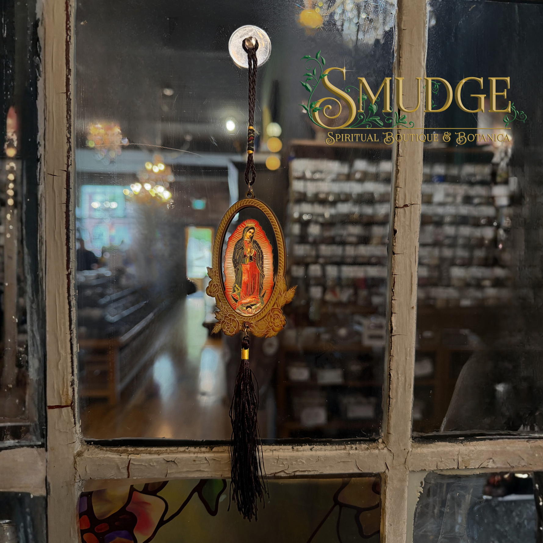 A religious hanging ornament featuring a small oval image of Our Lady of Guadalupe with gold-toned decorative details and a long black tassel, suspended in front of a shop window. The window has the words 'SMUDGE Spiritual Boutique & Botany' printed on it.