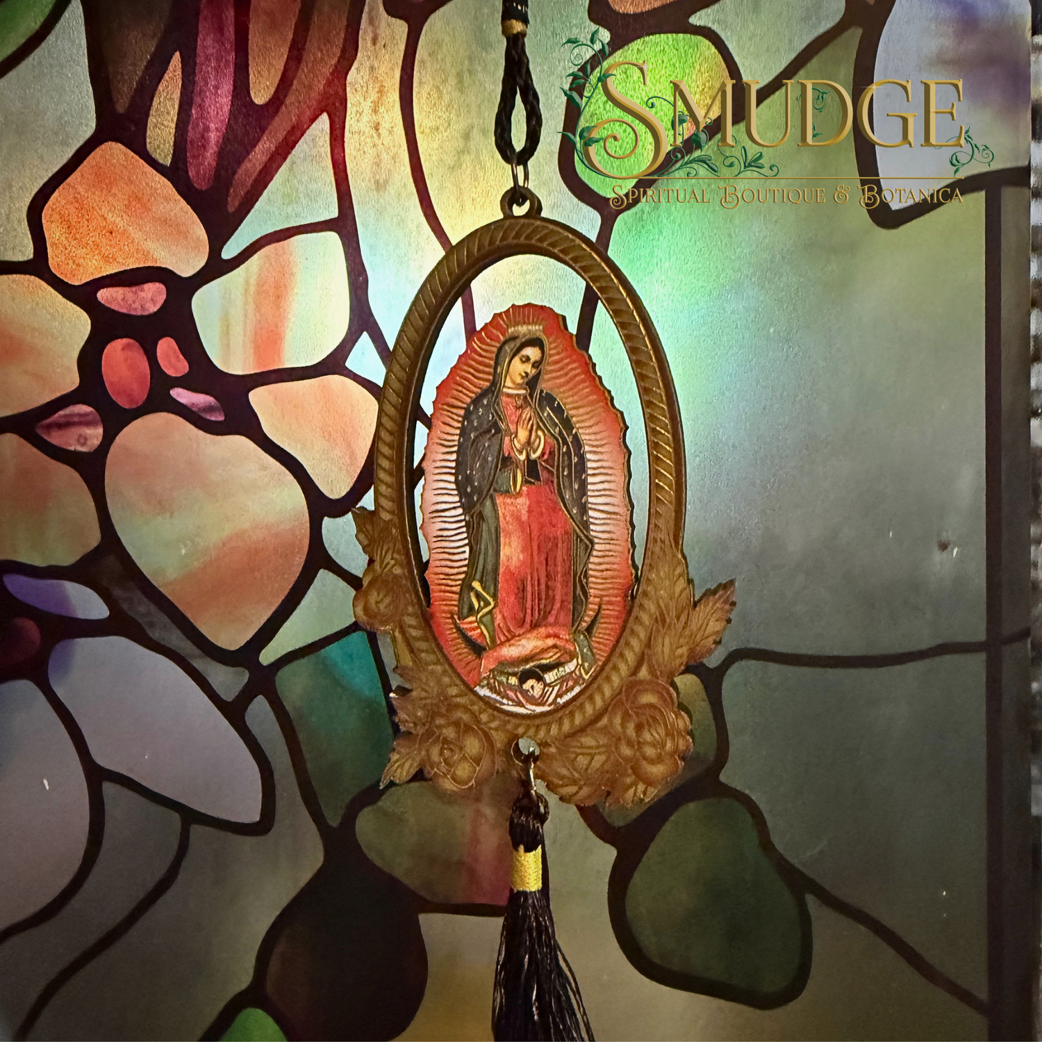 A stained glass window featuring Our Lady of Guadalupe hangs in front of a colorful stained glass panel. A gold-toned oval religious plaque with a black tassel is centered in the foreground.