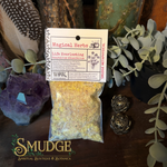 A clear plastic bag filled with yellow-green powdered herbal blend sits on a dark wooden surface, attached to a white paper label that reads 'Magical Herbs' and 'Life Everlasting'. The scene is styled with botanical props including eucalyptus leaves, a purple crystal cluster, and two decorative metal ornaments.