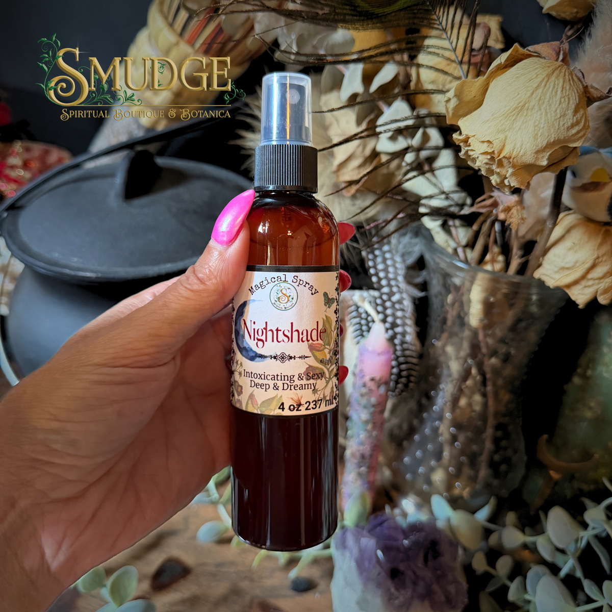 A hand with pink nail polish holds a 4 oz amber glass spray bottle of Smudge Spiritual Botanica 'Nightshade' essential oil spray. The background is a styled flat-lay of dried flowers, crystals, and a black teapot.