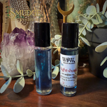 Two perfume oil rollerball bottles on a wooden surface, styled with crystals, greenery, and a gold crescent moon ornament. The left bottle contains a shimmering blue liquid, and the right bottle has a white label reading Witch of Wildervill Nightshade.