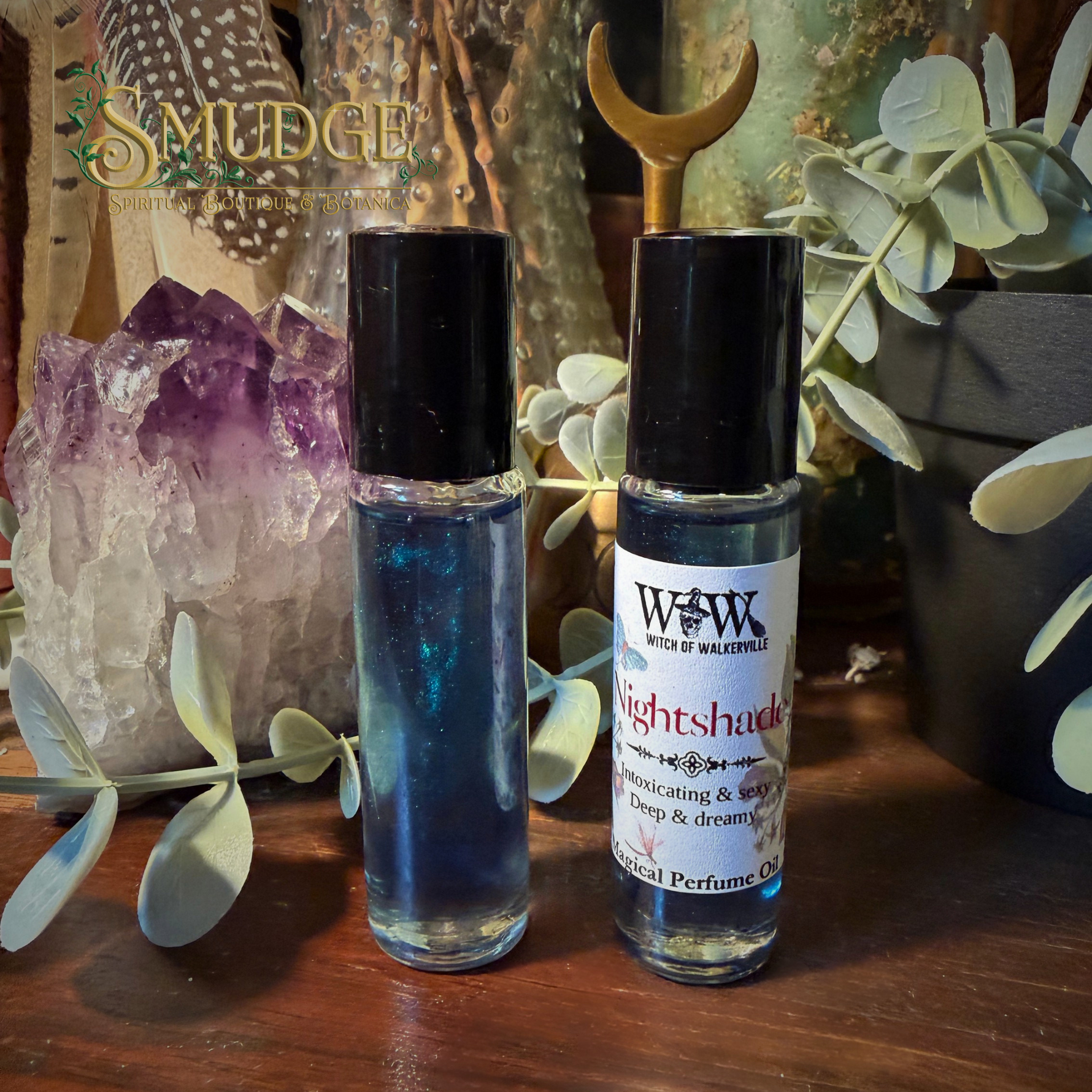Two perfume oil rollerball bottles on a wooden surface, styled with crystals, greenery, and a gold crescent moon ornament. The left bottle contains a shimmering blue liquid, and the right bottle has a white label reading Witch of Wildervill Nightshade.